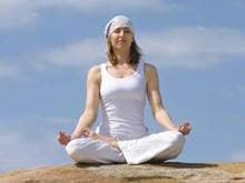 A woman in white clothes and a bandana meditating on a rock against the blue sky.