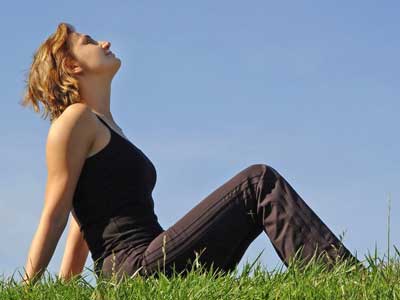 A woman sitting on a green grass field, looking up into a clear blue sky.
