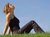 A woman sitting on a green grass field, looking up into a clear blue sky.